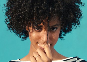 Woman wearing a black and white striped shirt and two rings with coily hair pulls a single lock down in front of her on a teal background.