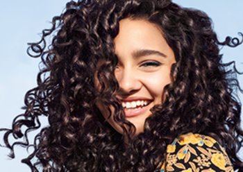 Woman with dark curly hair wearing a yellow flower-print shirt laughs while looking at the camera with a blue sky behind her.