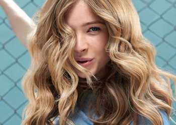 Woman with blonde, wavy hair laughs infront of a blue background and a chain link fence.
